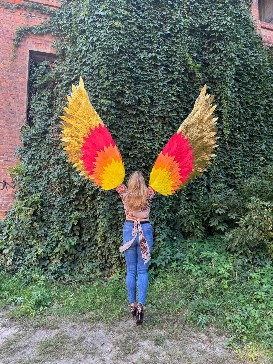 May include: A person wearing large, colorful angel wings against a backdrop of lush green foliage. The wings feature a gradient of gold, red, and yellow hues, creating a striking visual effect. The wings are the focal point, suggesting a theme of fantasy or costume.