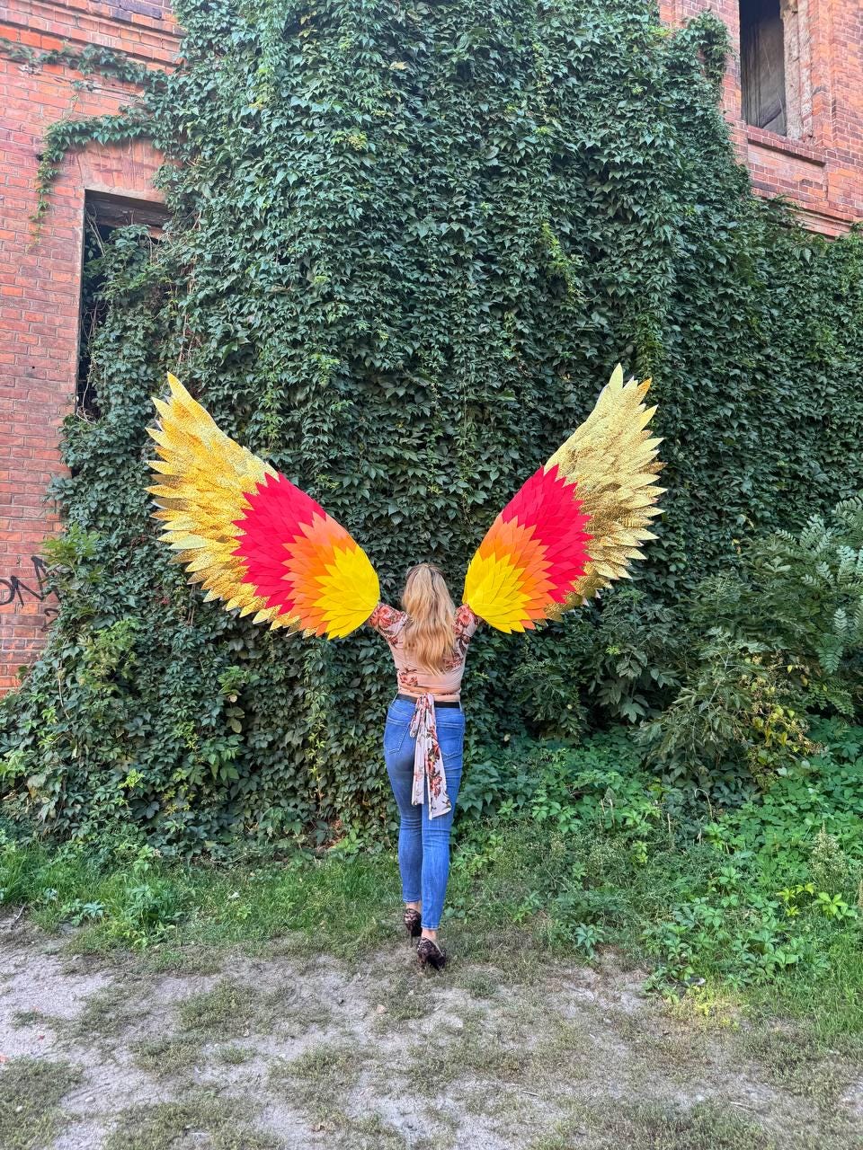 May include: A person wearing large, colorful wings with gold, yellow, red, and orange hues, standing in front of a wall covered in green foliage. The wings are spread wide, creating a striking visual effect. The person is wearing a floral top and blue jeans.