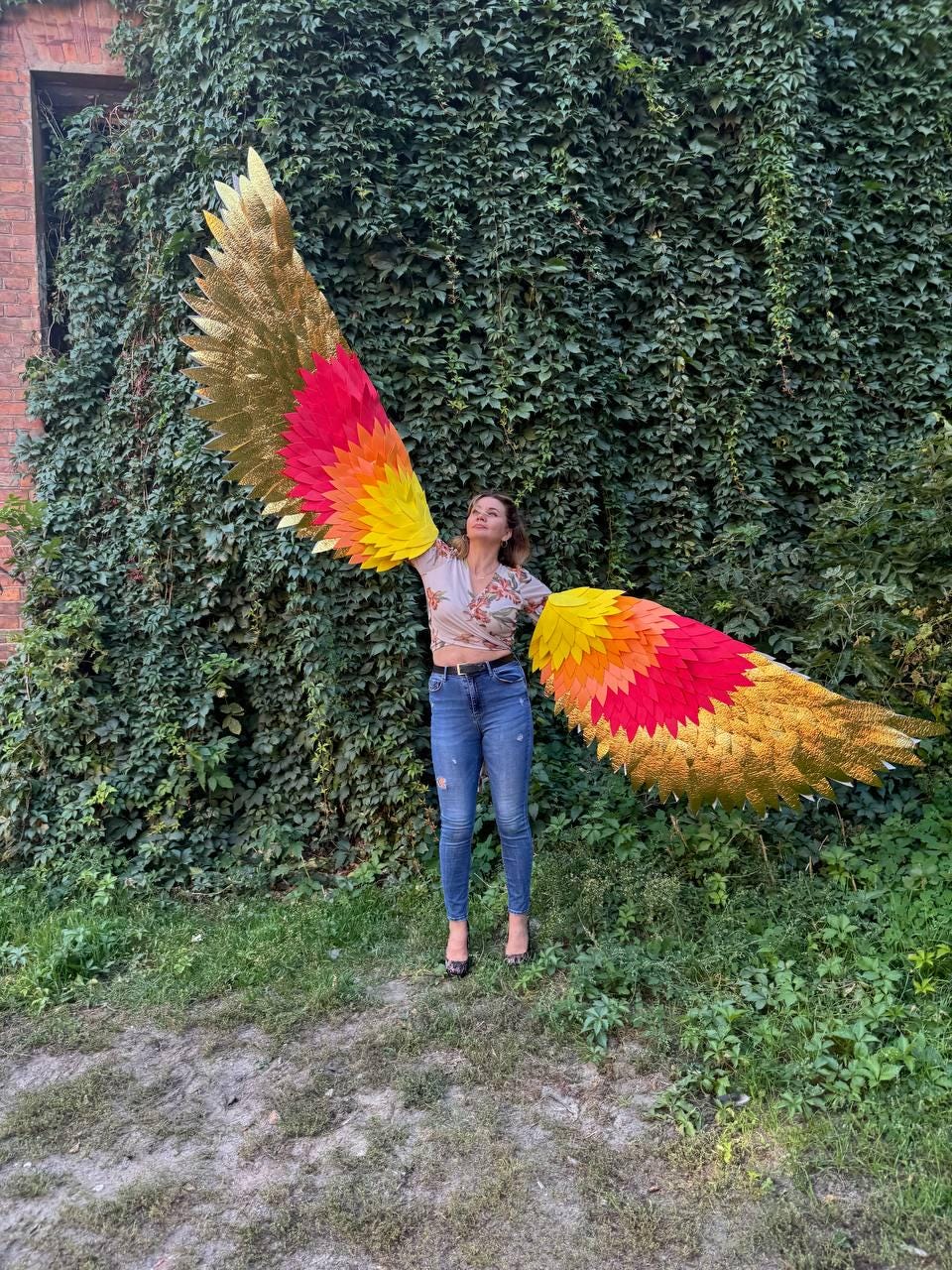 May include: Large, colorful wings with gold, red, orange, and yellow feathers. The wings are spread wide against a backdrop of green foliage. The wings appear to be made of layered materials.