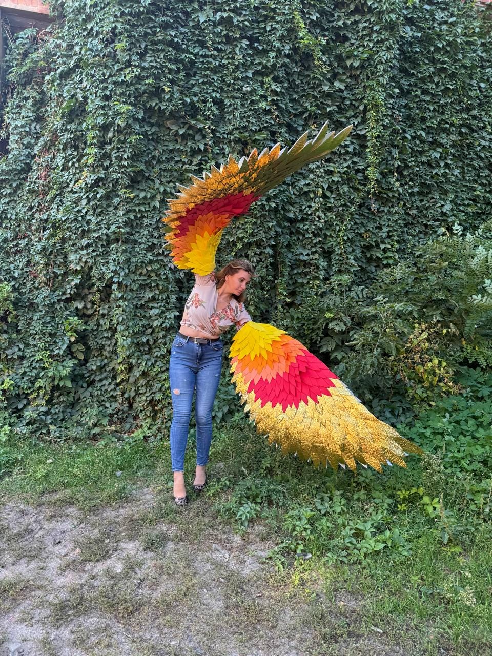 May include: Large, colorful, handmade wings with a gradient of yellow, orange, and red feathers. The wings are displayed outdoors against a backdrop of green foliage. The wings are designed to be worn.
