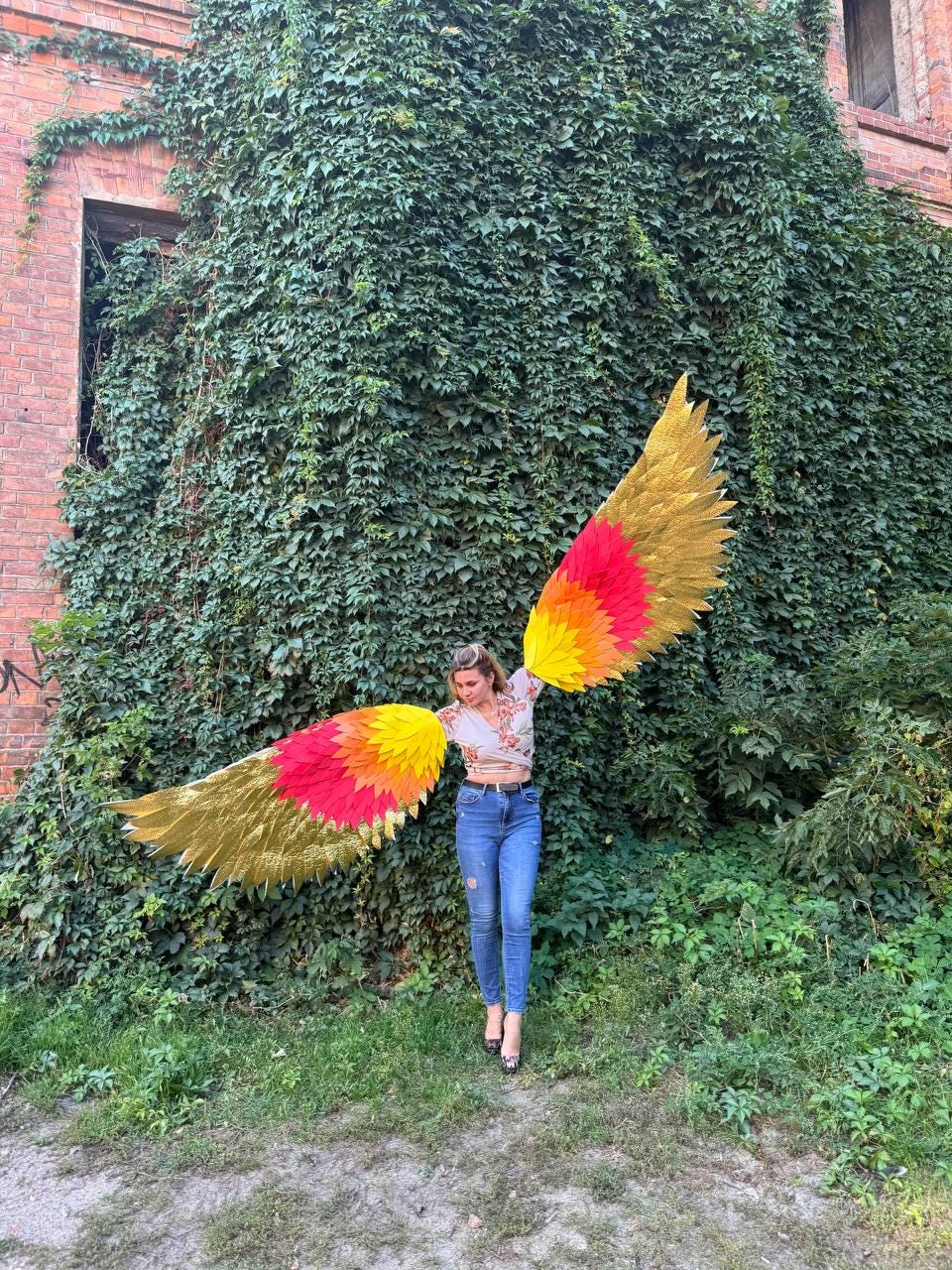 May include: A person wearing large, colorful wings with gold, red, and yellow hues, standing in front of a wall covered in green foliage. The wings are spread wide, showcasing their vibrant design.