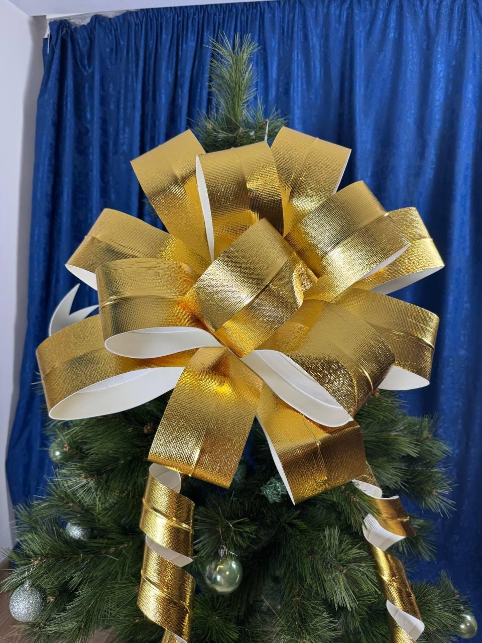 May include: A large, gold-colored bow made of metallic ribbon, with white undersides, sits atop a green Christmas tree. The bow is the focal point, with its multiple loops and layers. The tree is decorated with small, silver and green ornaments.
