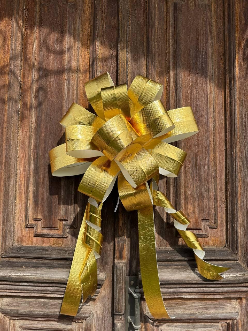 May include: A large, decorative gold bow with long, curling streamers. The bow is made of shiny, metallic material and is attached to a dark wooden door. The bow's loops and streamers create a festive and elegant appearance.