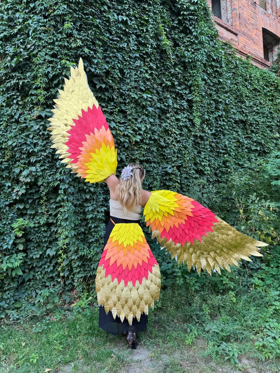 May include: A person wearing a colorful costume with large, feathered wings. The wings and skirt feature a gradient of yellow, orange, and red, with gold accents. The costume is set against a backdrop of green foliage and a brick building.