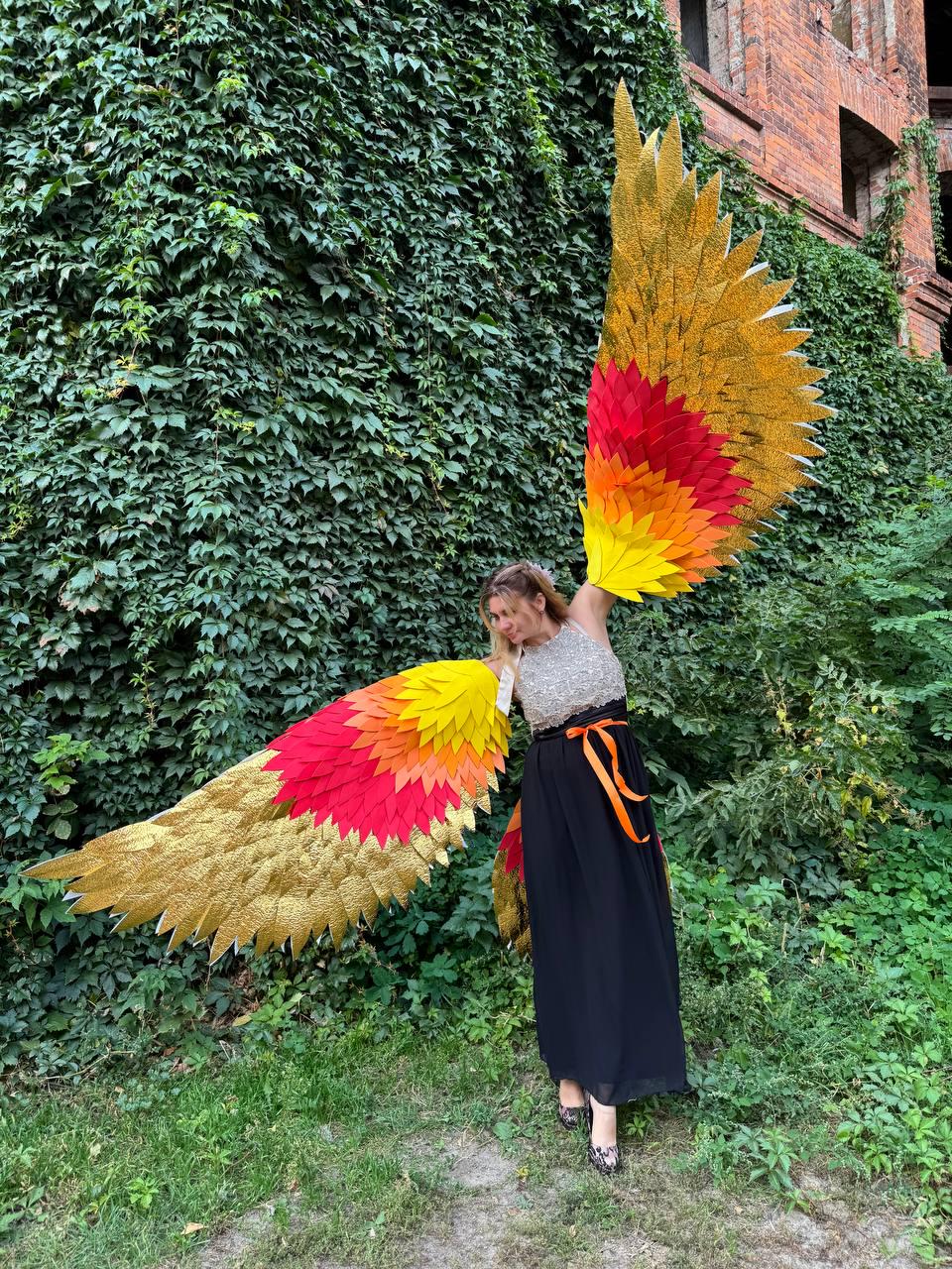 May include: A person wearing large, colorful, feathered wings. The wings feature gold, red, orange, and yellow hues. The person is wearing a silver top and a black skirt with an orange ribbon. The background is a wall of green foliage.