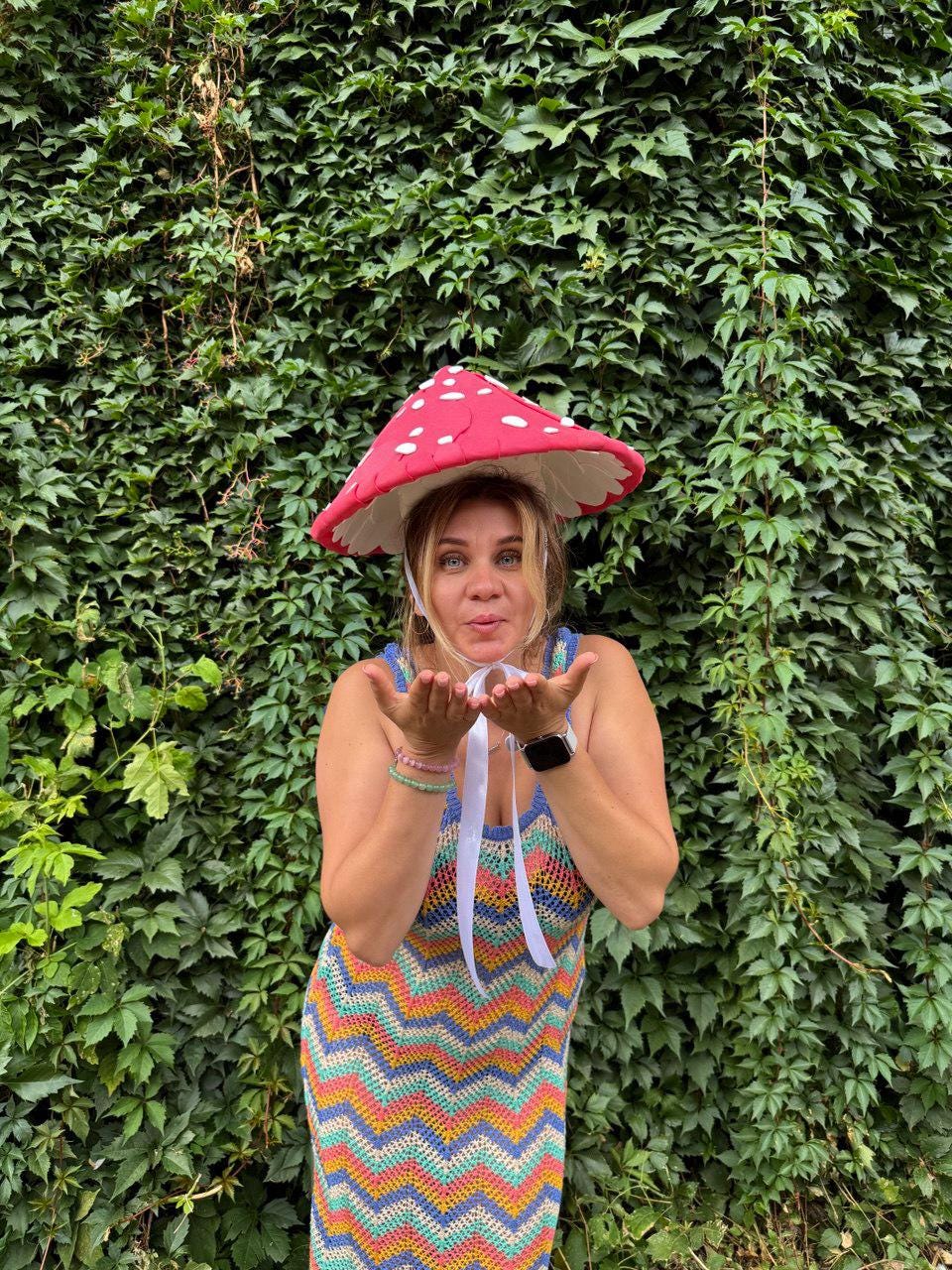 May include: A woman wearing a red mushroom hat with white polka dots and a colorful chevron-patterned dress. The hat has a white trim and is tied with a white ribbon. The dress features a mix of blue, orange, yellow, and green colors.