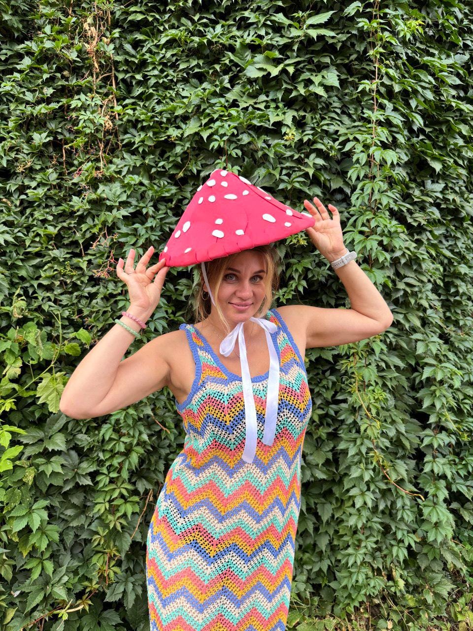 May include: A woman wearing a red mushroom-shaped hat with white spots and a colorful crochet dress. The dress features a chevron pattern in shades of blue, yellow, pink, and green. White ribbons are tied at the neckline. The background is a wall of green leaves.