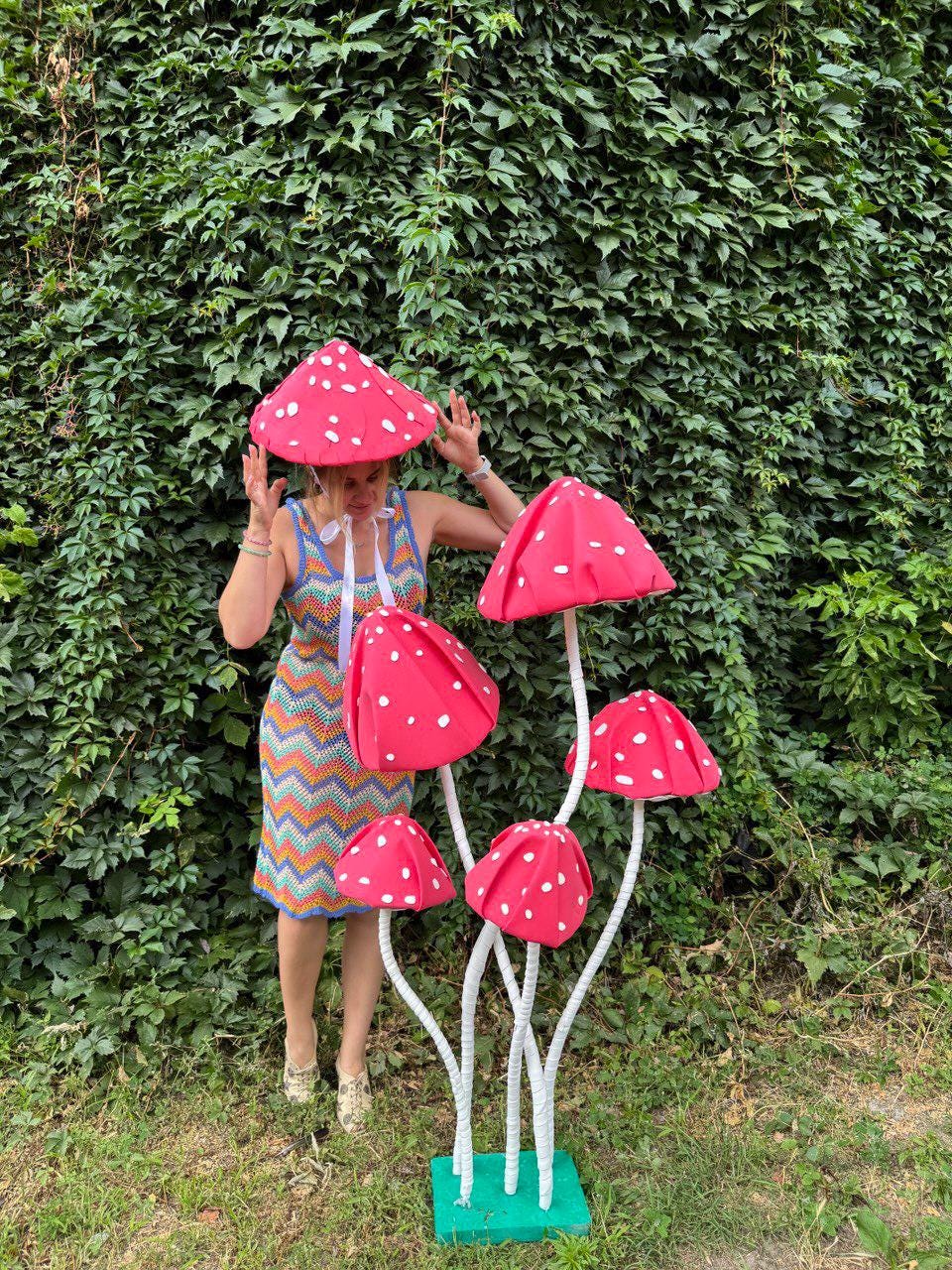May include: A whimsical display featuring large, red mushroom decorations with white polka dots. The mushrooms are supported by white stems and a green base. A person is wearing a matching mushroom hat and a colorful dress.