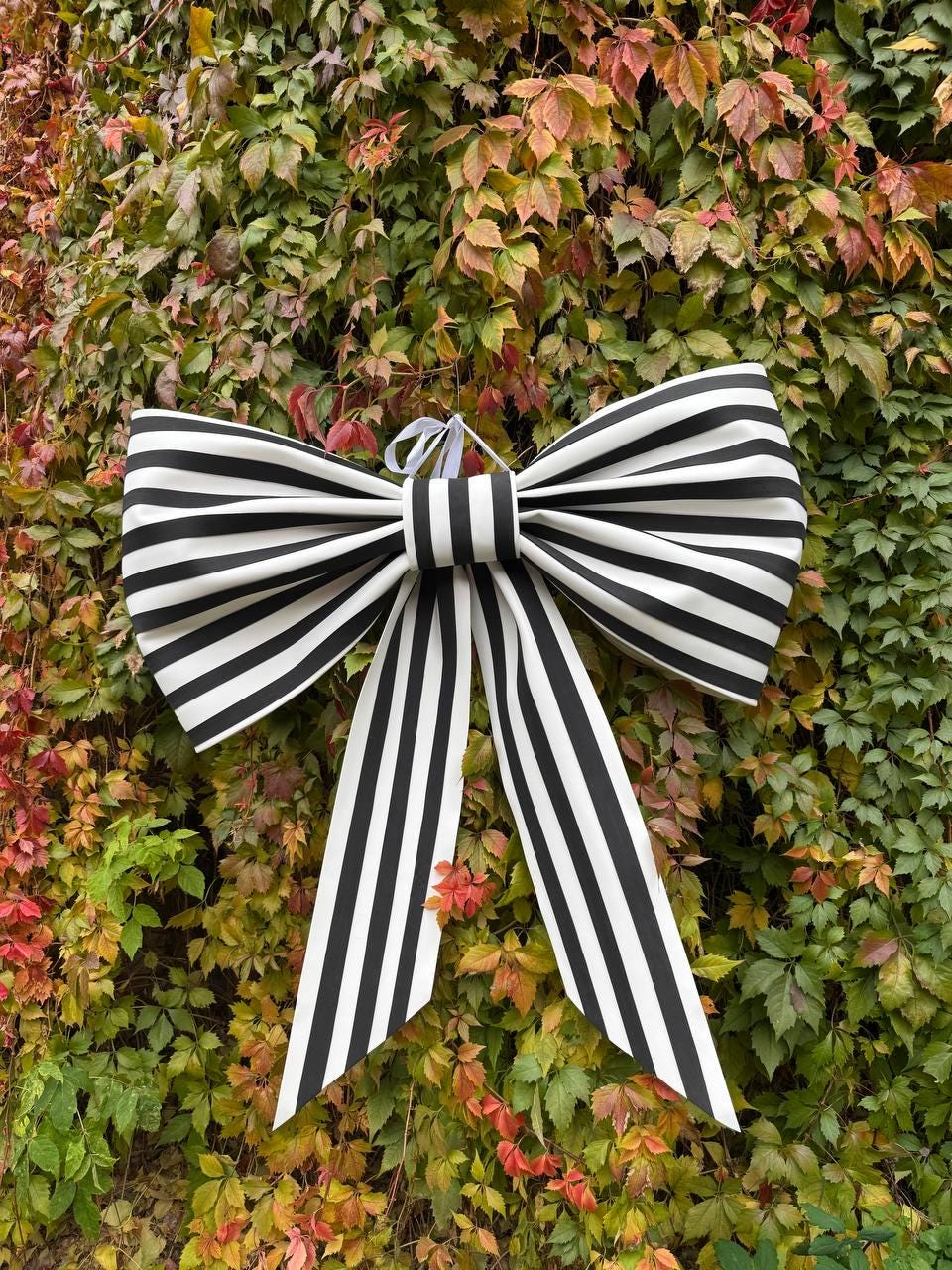 May include: A large decorative bow with black and white stripes, hanging against a backdrop of green and red foliage. The bow has long, striped tails and a central knot. The bow is likely used for decoration.