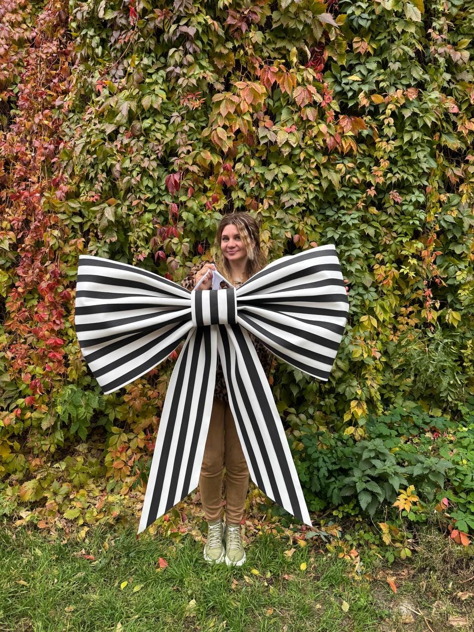 May include: A large, decorative bow with black and white stripes. The bow is held in front of a person standing in front of a wall of colorful autumn foliage. The bow's design is bold and eye-catching, suitable for events or decor.