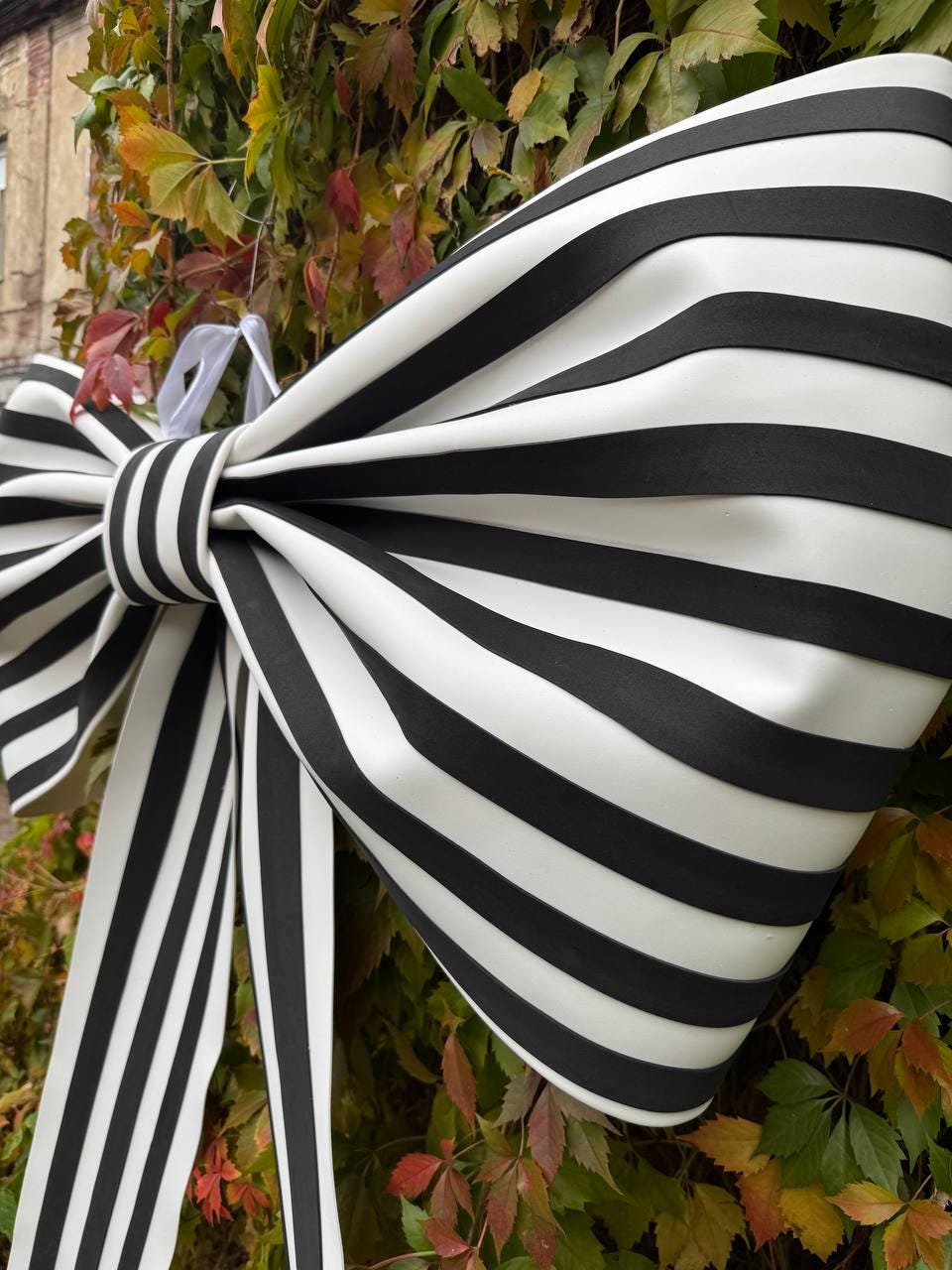 May include: A large, decorative bow with bold black and white stripes. The bow is made of a smooth material and has long, flowing ribbons. The bow is set against a backdrop of green and red foliage.