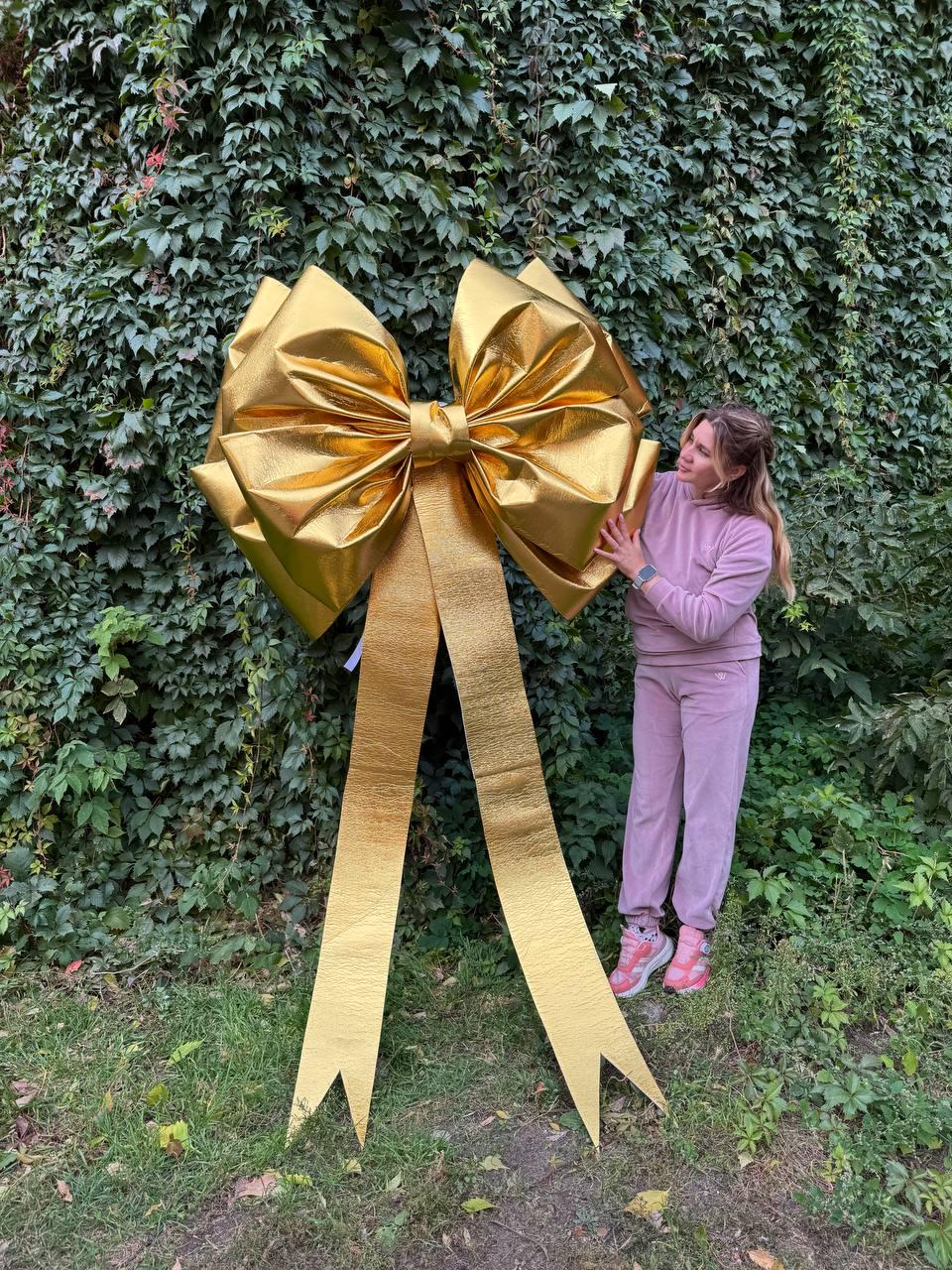May include: A large, golden bow with long, flowing ribbons. The bow is made of a shiny, metallic material and is positioned in front of a wall of green foliage. The bow is a decorative element.