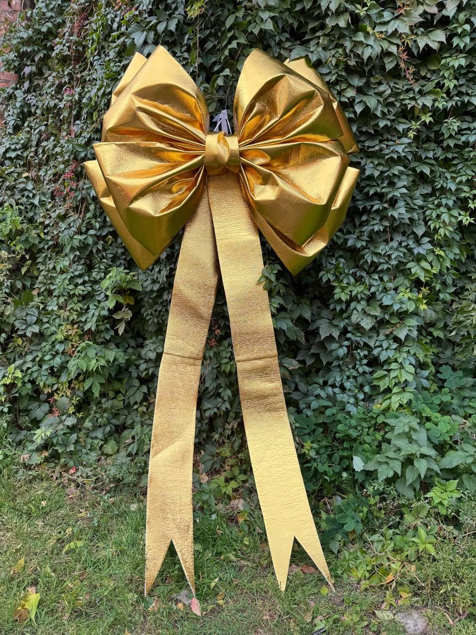 May include: A large, gold-colored bow with long, pointed streamers. The bow is made of a shiny, textured material and is set against a backdrop of green foliage and grass. The bow is likely used for decoration.