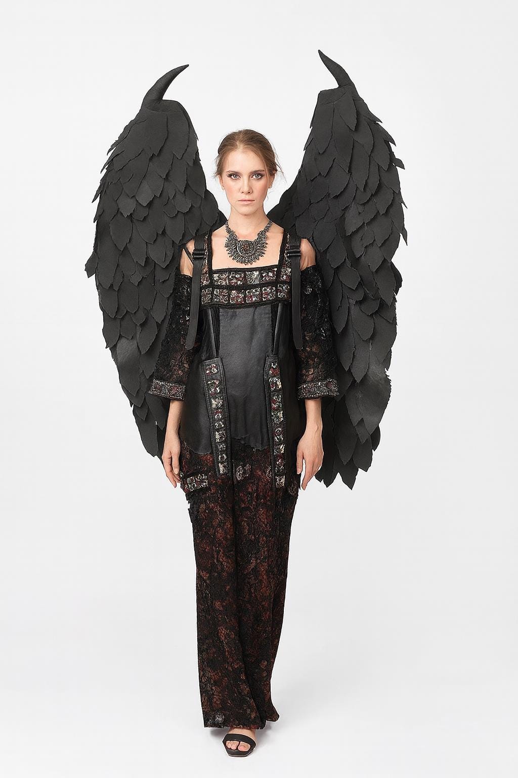 May include: A woman wearing a black gothic-style outfit with large black feathered wings. The dress features a leather bodice, lace sleeves, and a patterned skirt. A silver necklace completes the look. The wings are attached to the back of the outfit.
