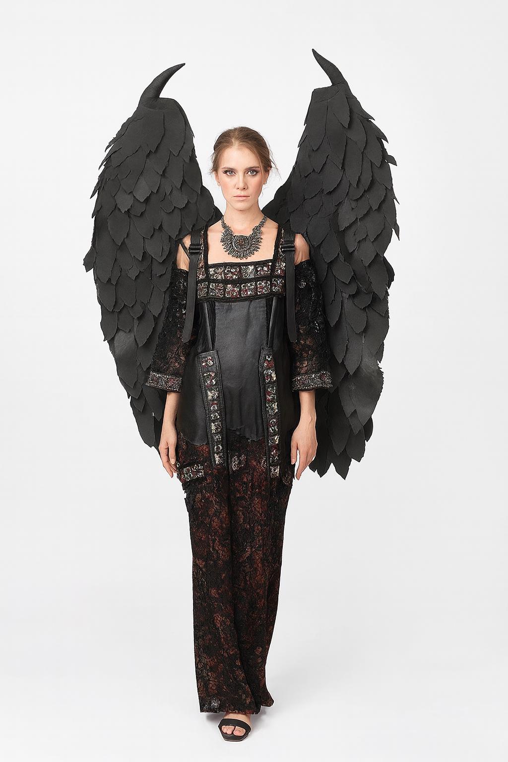 May include: A woman wearing a black gothic-style outfit with large black feathered wings. The dress features a leather bodice, lace sleeves, and a patterned skirt. A silver necklace completes the look. The wings are attached to the back of the outfit.