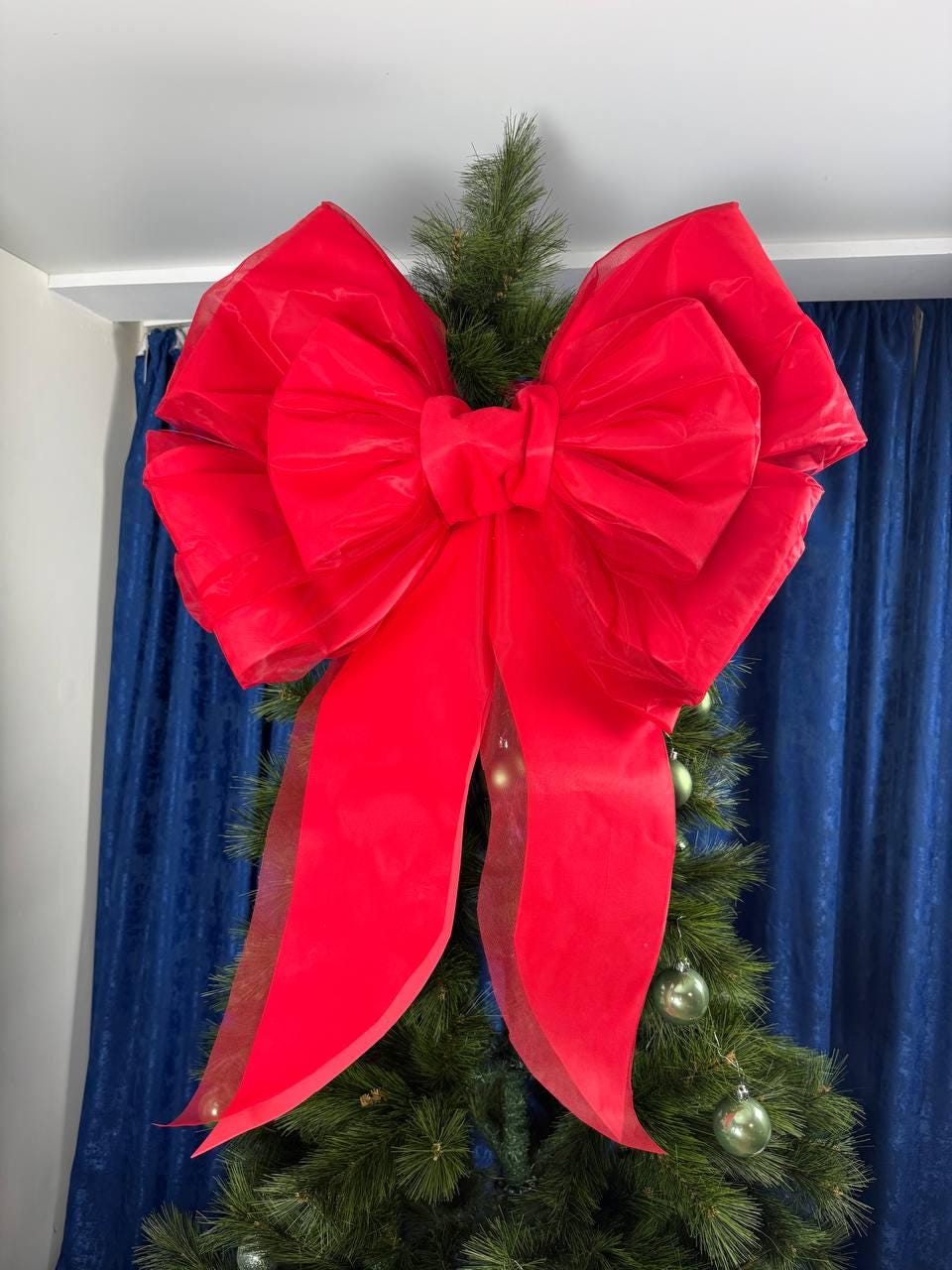 May include: A large, vibrant red bow adorns the top of a Christmas tree. The bow is made of a sheer, flowing material, with long, elegant streamers. The tree is decorated with green ornaments, set against a blue curtain backdrop.