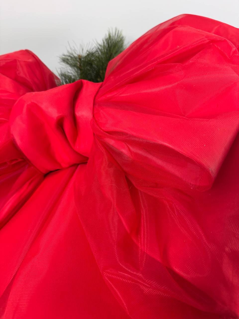 May include: A large, vibrant red bow made of a sheer, flowing fabric. The bow is intricately tied, with multiple loops and folds, and has a few sprigs of green pine needles peeking out from behind it. The bow is a festive decoration.