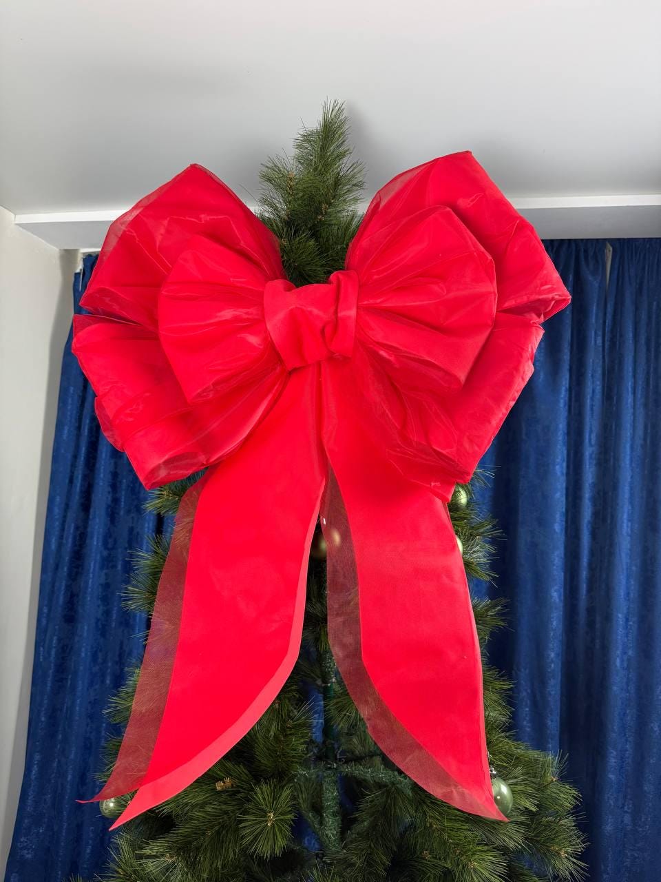 May include: A large, vibrant red bow made of sheer fabric, displayed on a Christmas tree. The bow has multiple layers and long, flowing tails. The tree is decorated with green ornaments and set against a blue curtain backdrop.