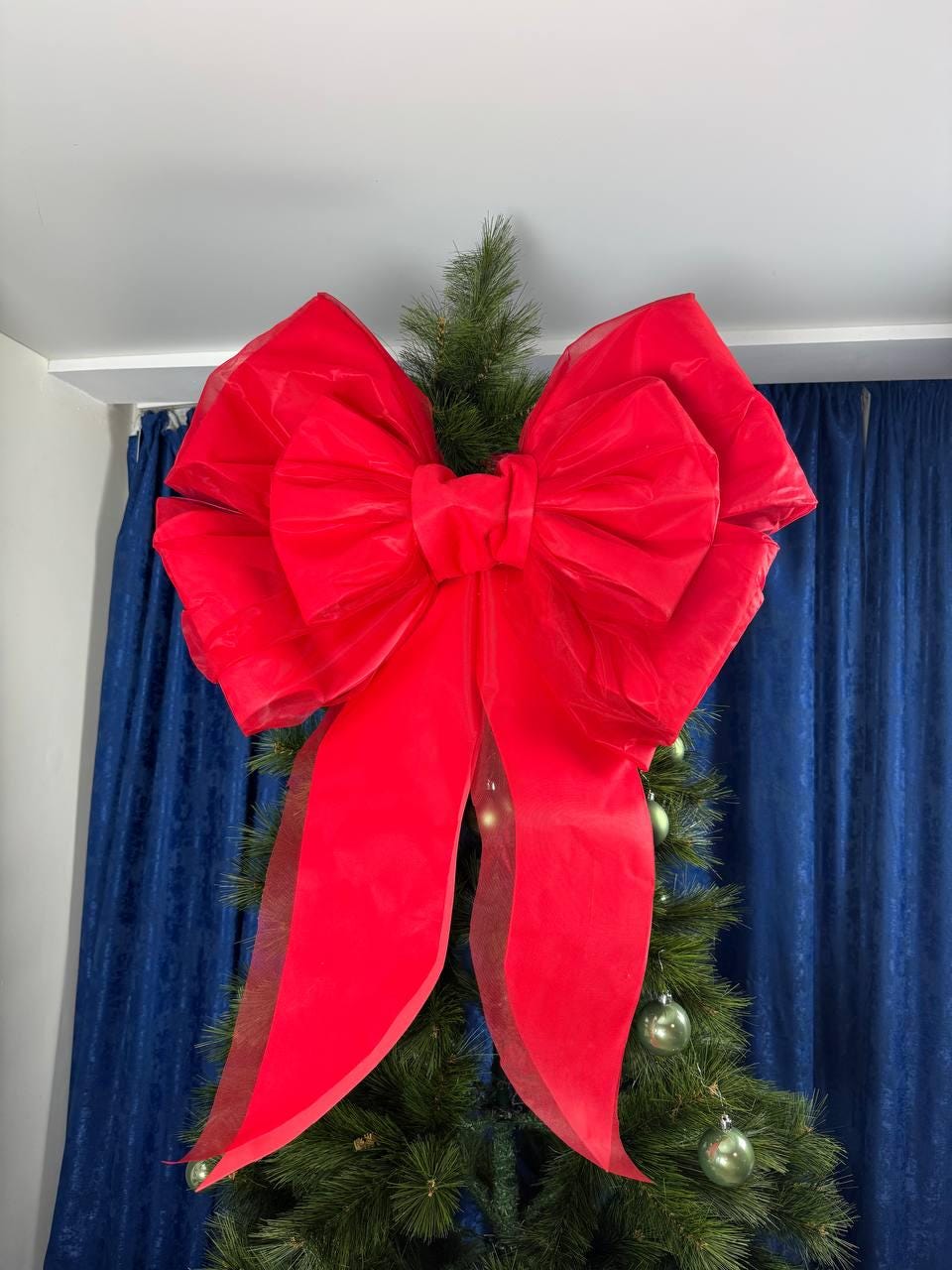 May include: A large, vibrant red bow adorns the top of a Christmas tree. The bow is made of a sheer, flowing fabric, with long, elegant streamers. The tree is decorated with green ornaments, set against a backdrop of blue curtains.