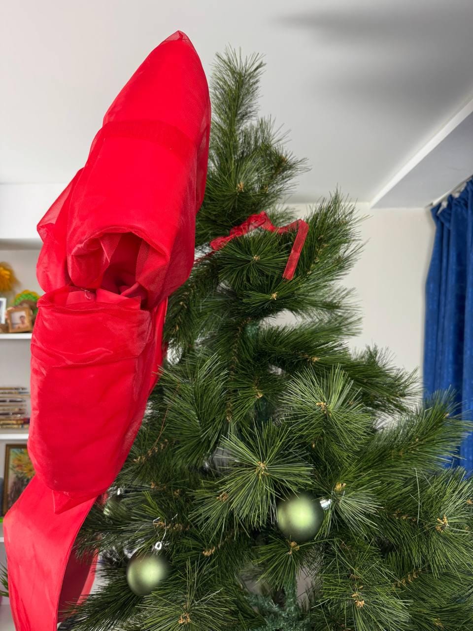 May include: A decorated Christmas tree with a large, bright red bow at the top. The tree is adorned with green ornaments and red ribbon. The tree's needles are a deep green color, creating a festive holiday display.