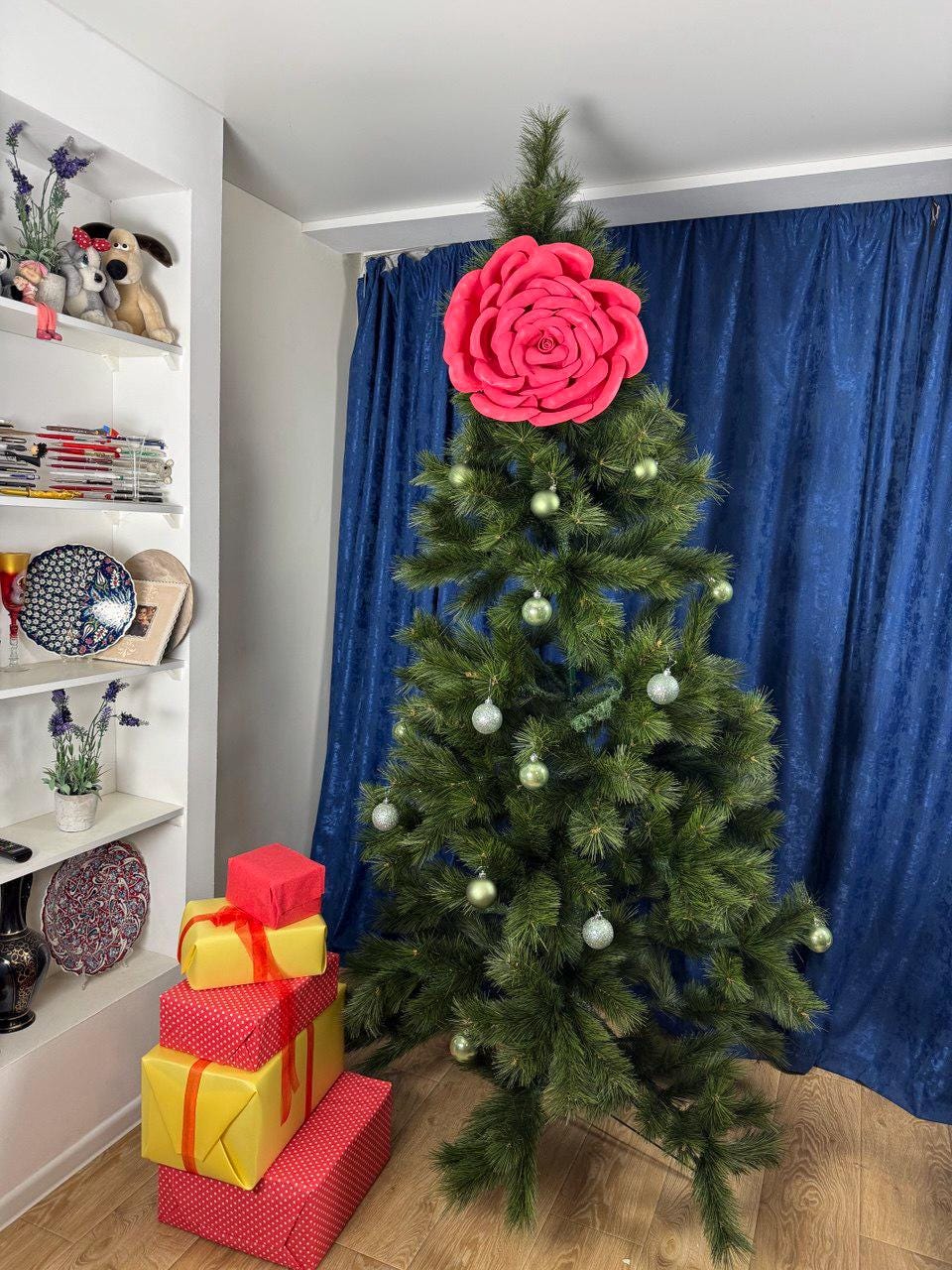 May include: A decorated Christmas tree with a large pink rose tree topper and silver ornaments. A stack of wrapped gifts in red and yellow sits at the base of the tree. The tree is in front of a blue curtain.