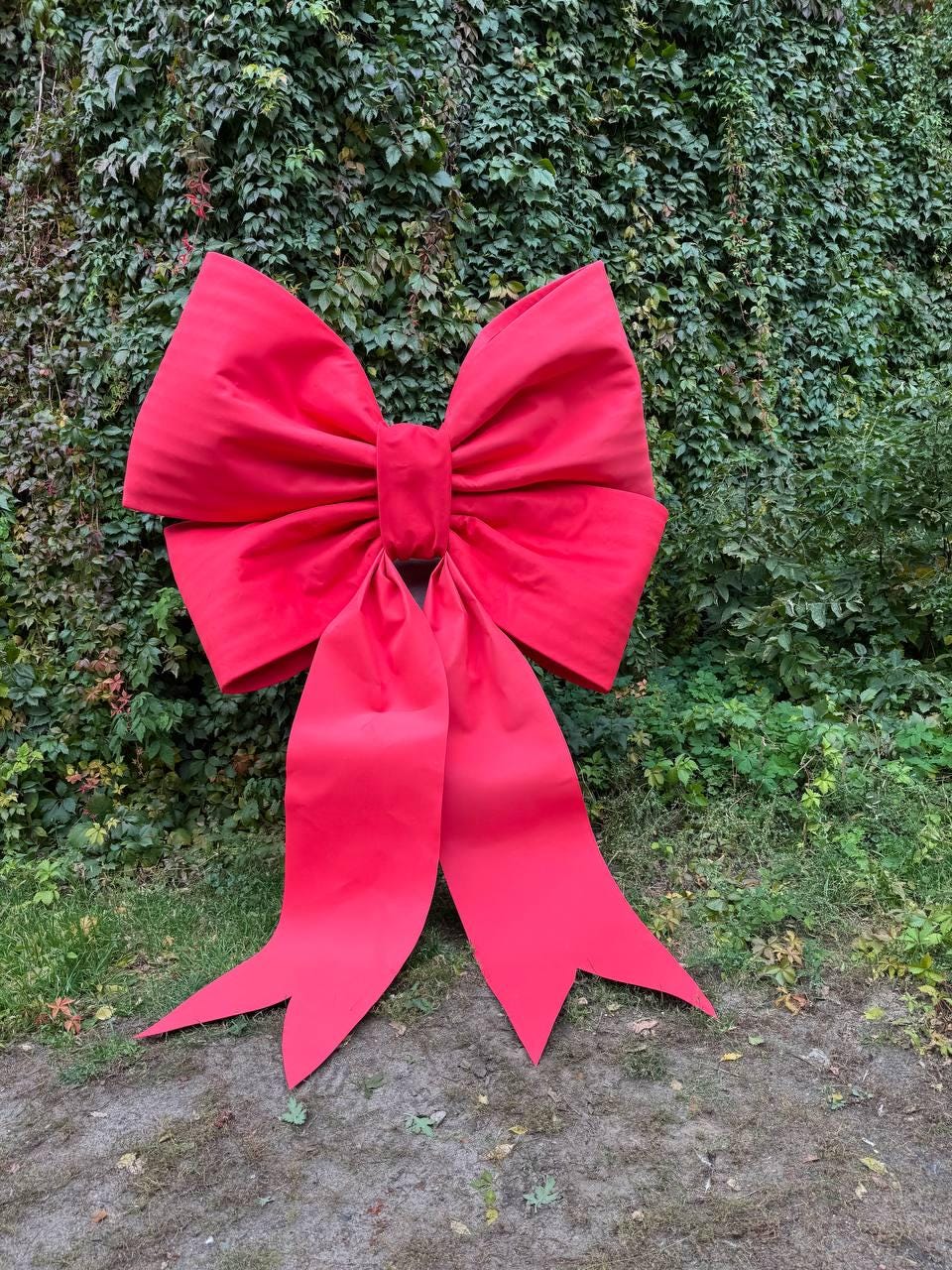May include: A large, vibrant red bow with long, pointed tails. The bow is made of a textured fabric and is positioned in front of a lush green wall of foliage. The bow is a festive decoration.