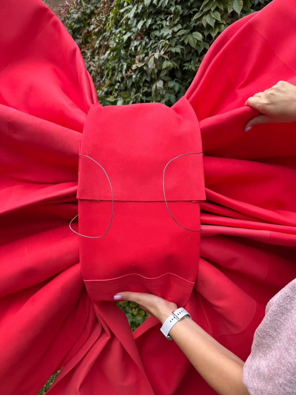 May include: A large, vibrant red bow made of fabric. The bow is the focal point, with its voluminous folds and central knot. The bow is held by hands, suggesting its size and scale. The background is a lush green foliage.