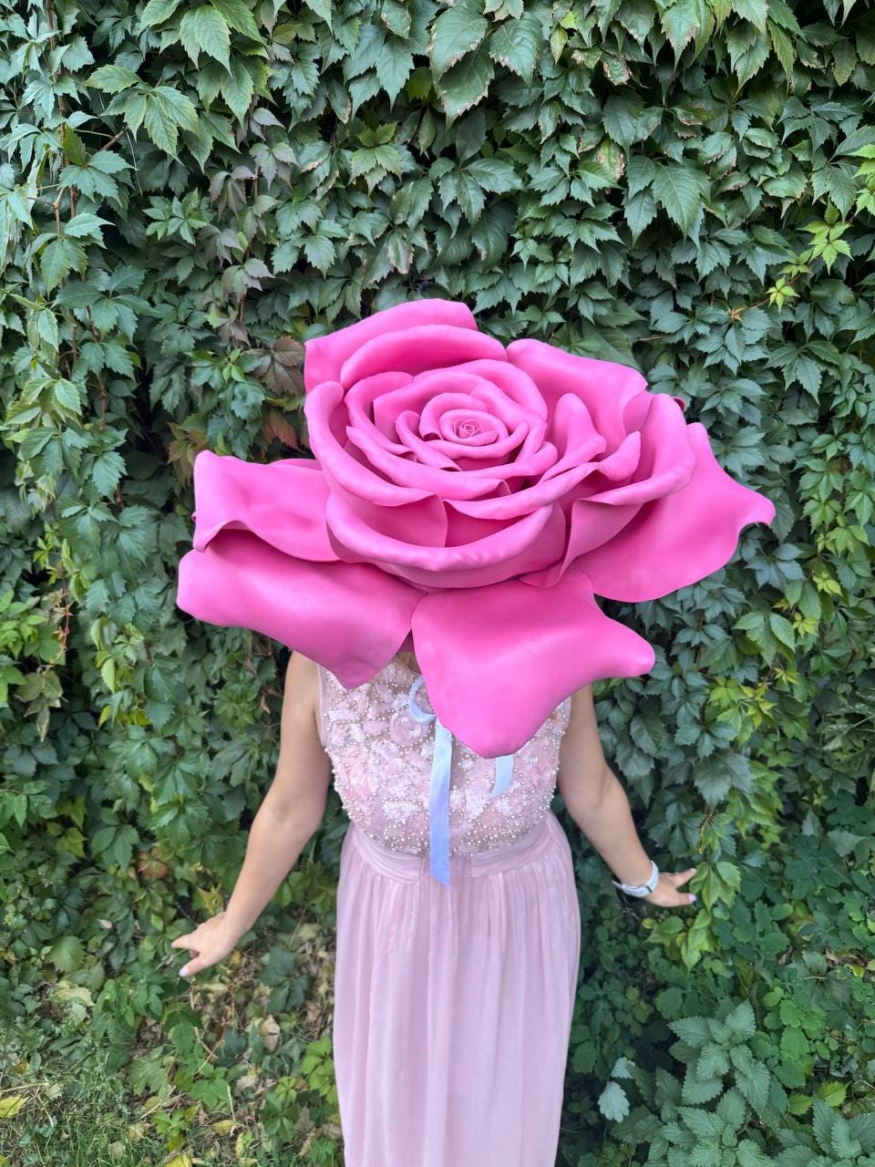 May include: A large, vibrant pink rose headpiece, worn by a person in a light pink dress. The rose is the focal point, with detailed petals. The background is a lush green wall of foliage.