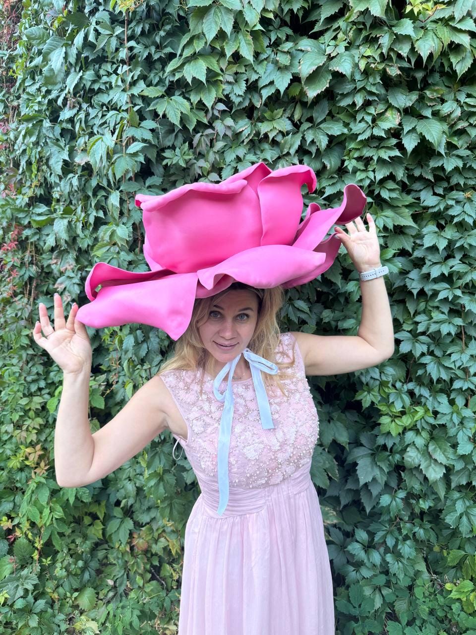 May include: A woman wearing a large, vibrant pink rose-shaped hat. She is dressed in a light pink, sleeveless dress with a beaded bodice and a light blue ribbon. The background is a wall of green leaves.