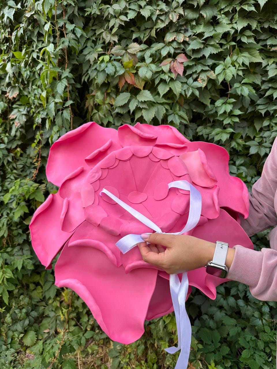 May include: A large, vibrant pink flower with layered petals and a scalloped center. White ribbon is attached to the back. The flower is held in front of a green leafy background.