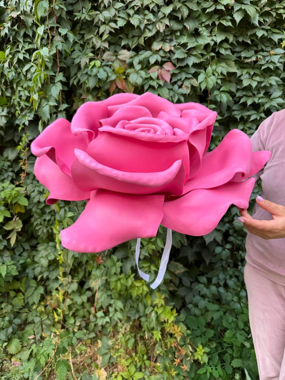 May include: A large, vibrant pink rose made of a soft material. The rose has multiple layers of petals and a white ribbon attached. The background is a lush green wall of leaves.