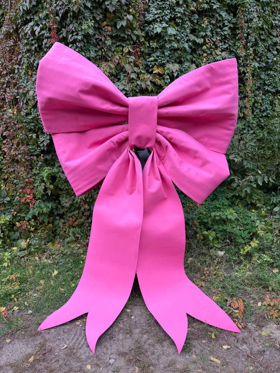 May include: A large, vibrant pink bow with long, flowing tails. The bow is made of a smooth, possibly satin-like material and is set against a backdrop of green foliage. The bow's size suggests it could be used for decoration.
