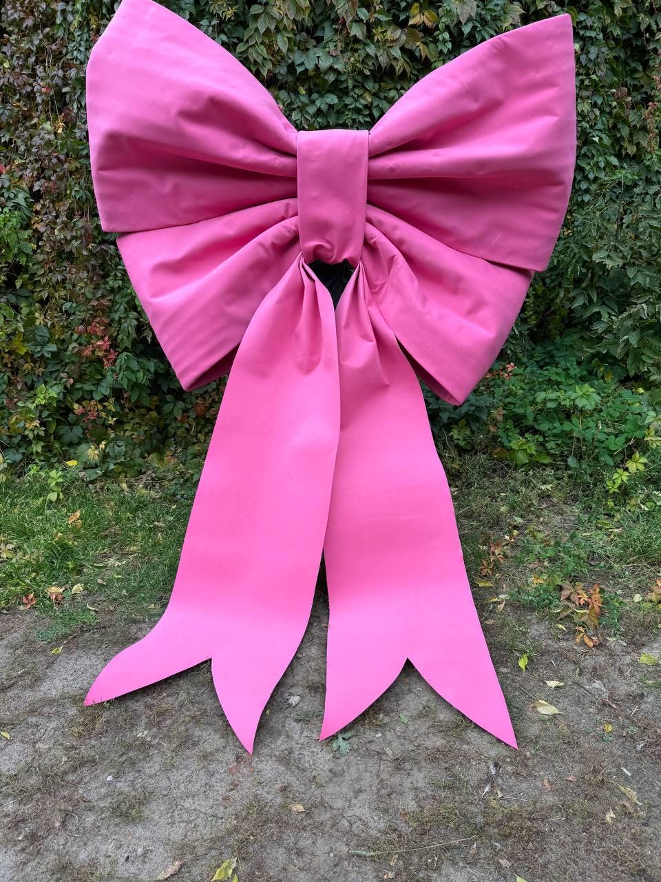 May include: A large, vibrant pink bow with long, flowing tails. The bow is made of a smooth, possibly fabric material, and is displayed outdoors against a backdrop of green foliage and grass. The bow's size suggests it could be used for decoration.