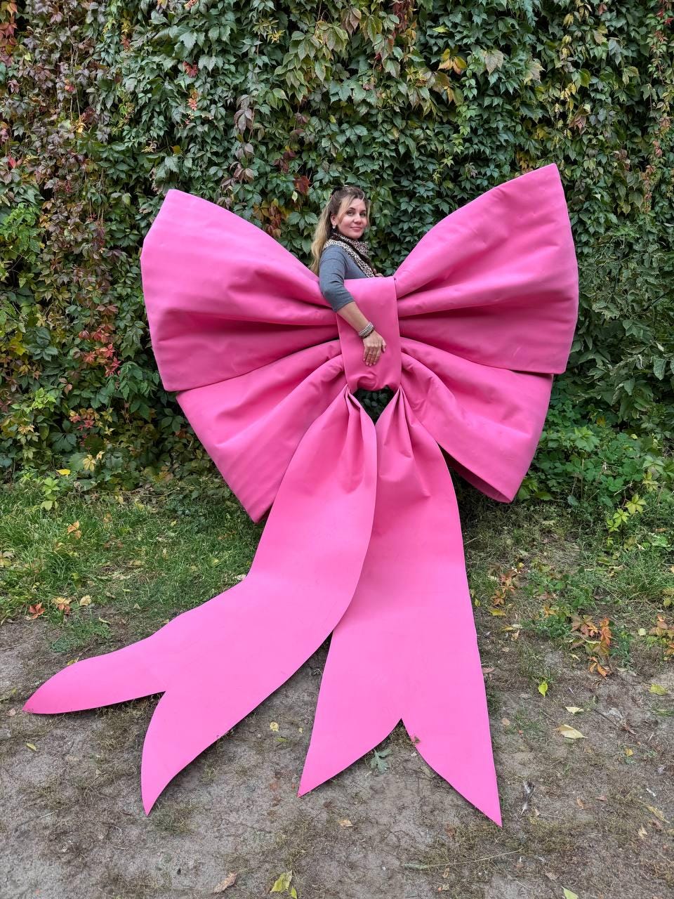 May include: A large, vibrant pink bow, standing upright on the grass. The bow is made of a smooth, possibly fabric material, with long, flowing tails. The bow is large enough for a person to stand behind it. Green foliage forms the background.