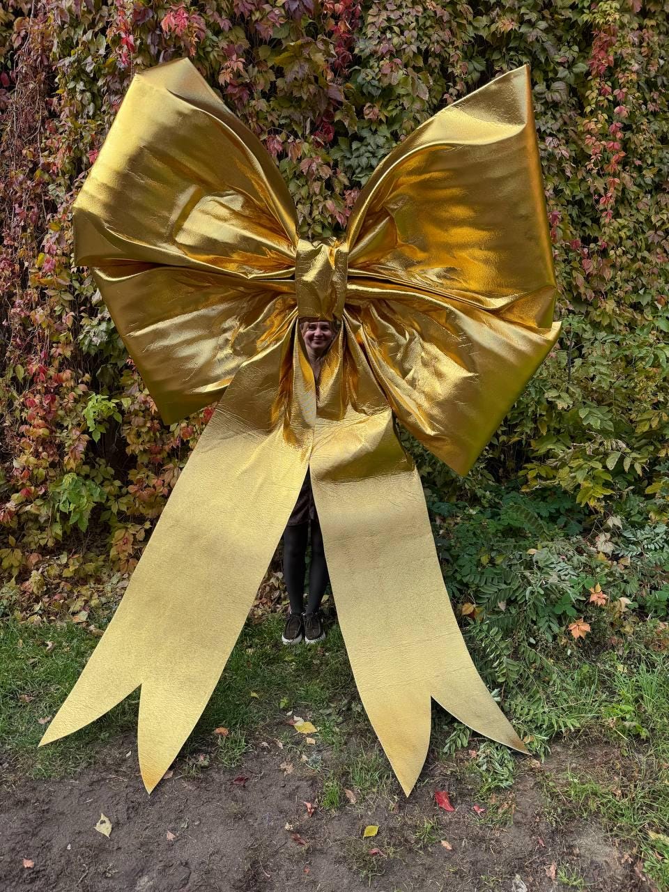 May include: A large, golden bow with long, pointed streamers. The bow is made of a shiny, metallic material and is positioned in front of a backdrop of green and red foliage. The bow is large enough for a person to stand behind it.