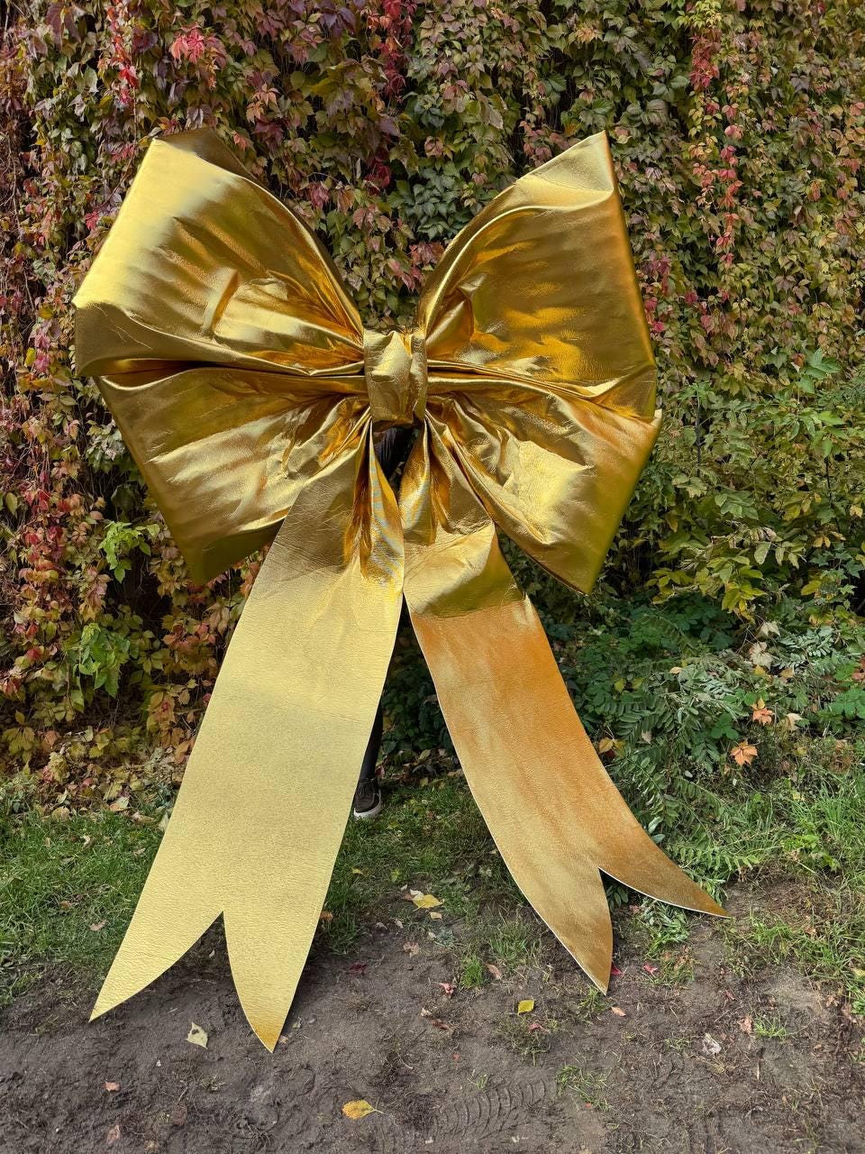 May include: A large, golden bow with long, flowing ribbons. The bow is made of a shiny, metallic material and is positioned outdoors against a backdrop of green and red foliage. The bow's size suggests it is a decorative element.