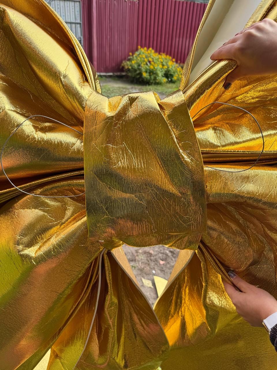 May include: A large, shiny gold bow made of textured fabric. The bow is intricately folded and has a metallic sheen. The bow is held by a person's hands, suggesting it is a decorative item, possibly for a gift or event.