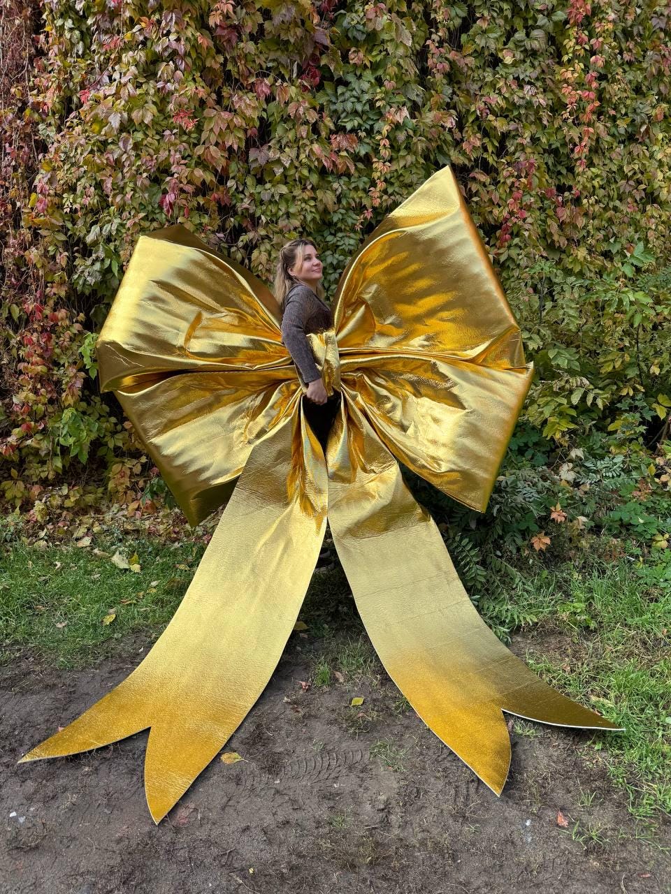 May include: A large, golden bow with long, flowing ribbons. The bow is made of a shiny, metallic material and is positioned outdoors against a backdrop of green and brown foliage. The bow's size suggests it is a decorative element.