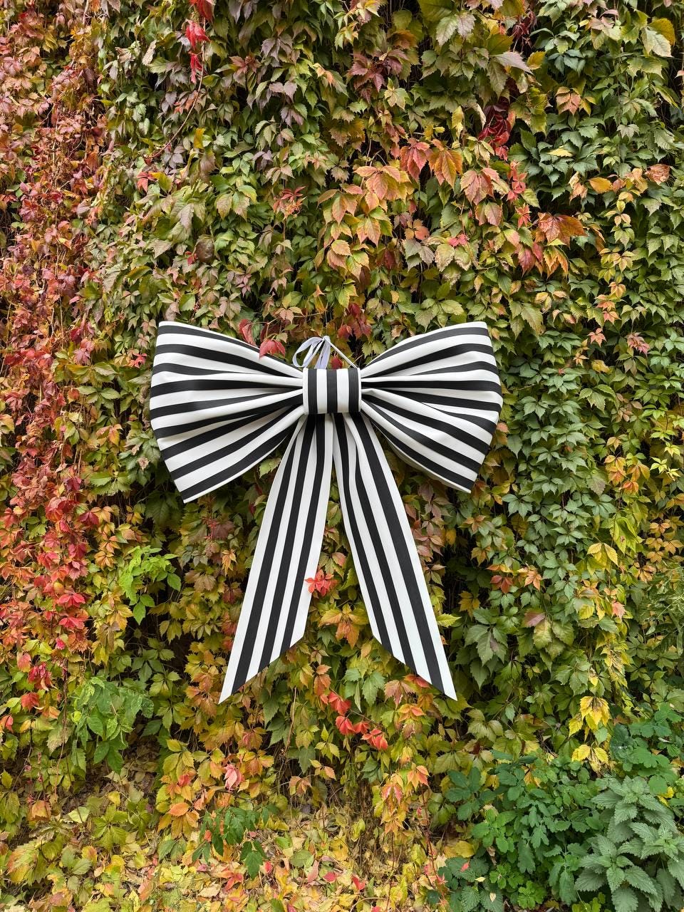 May include: A large decorative bow with black and white stripes hangs against a backdrop of colorful autumn foliage. The bow features a classic bow shape with long, striped tails, suitable for seasonal decor.