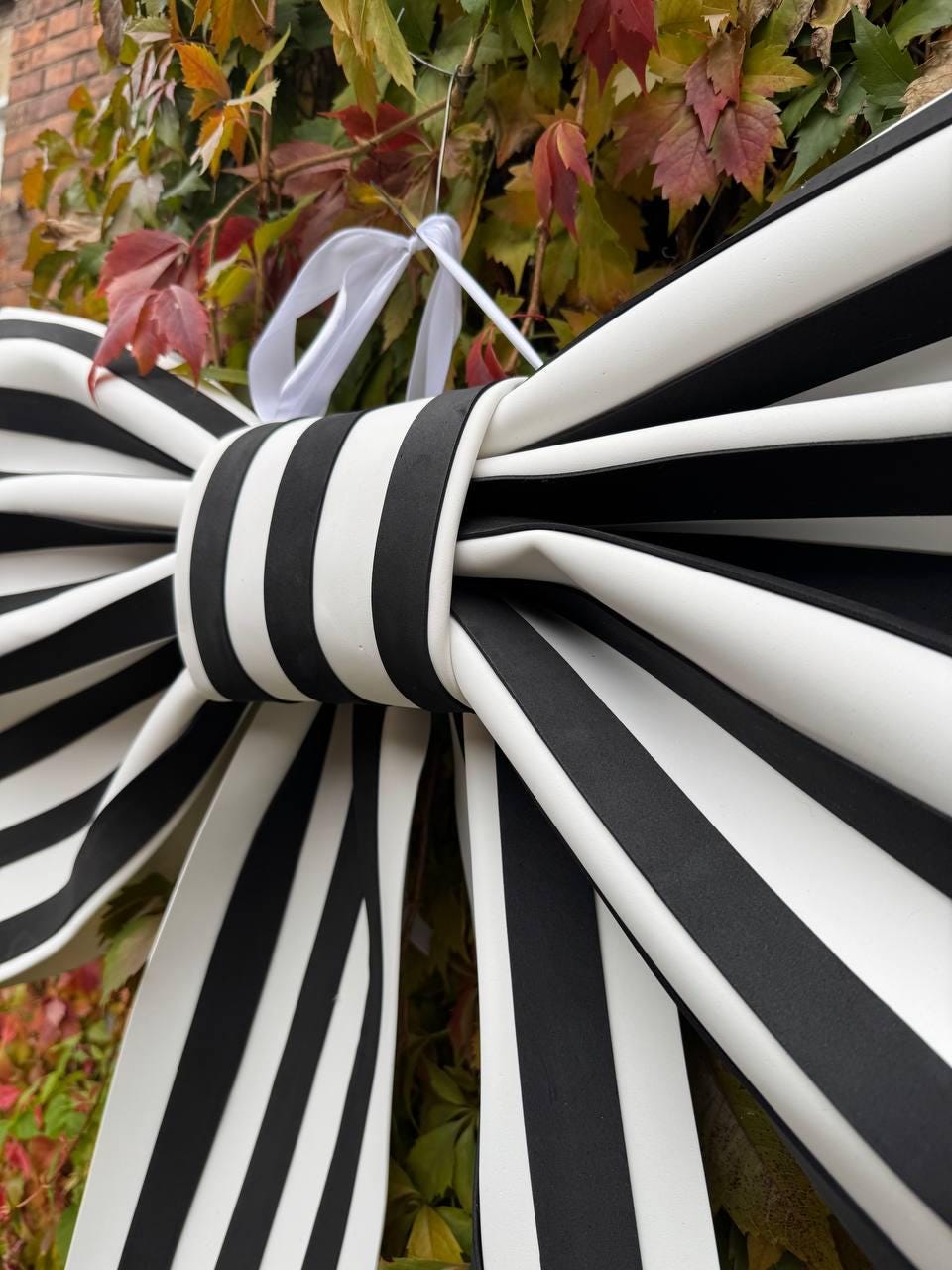 May include: A large decorative bow with bold black and white stripes. The bow is tied in the center and has long, flowing tails. A white ribbon is attached for hanging. The bow is set against a backdrop of colorful autumn foliage.