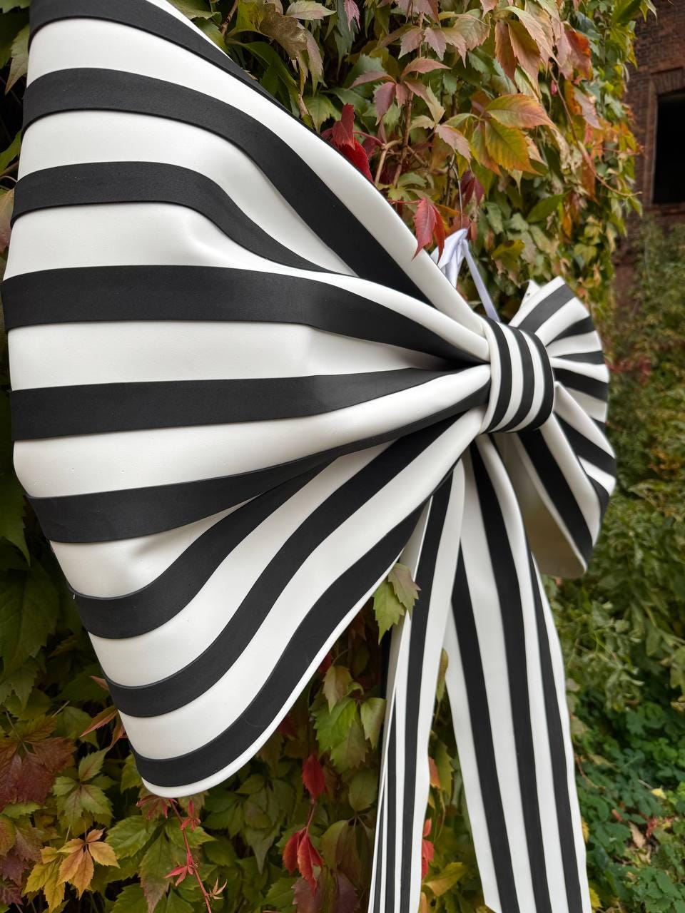 May include: A large, decorative bow with bold black and white stripes. The bow is tied in the center and has long, flowing tails. The background features green and red foliage, creating a striking contrast with the bow's design.