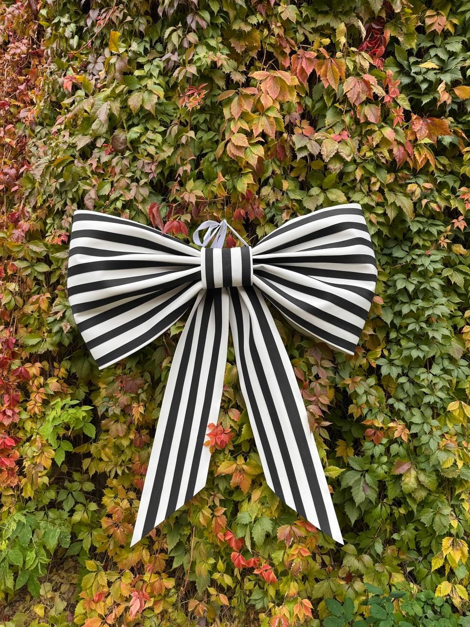 May include: A large decorative bow with a black and white striped pattern. The bow is made of fabric and has long, striped tails. It is hanging against a backdrop of colorful autumn foliage.