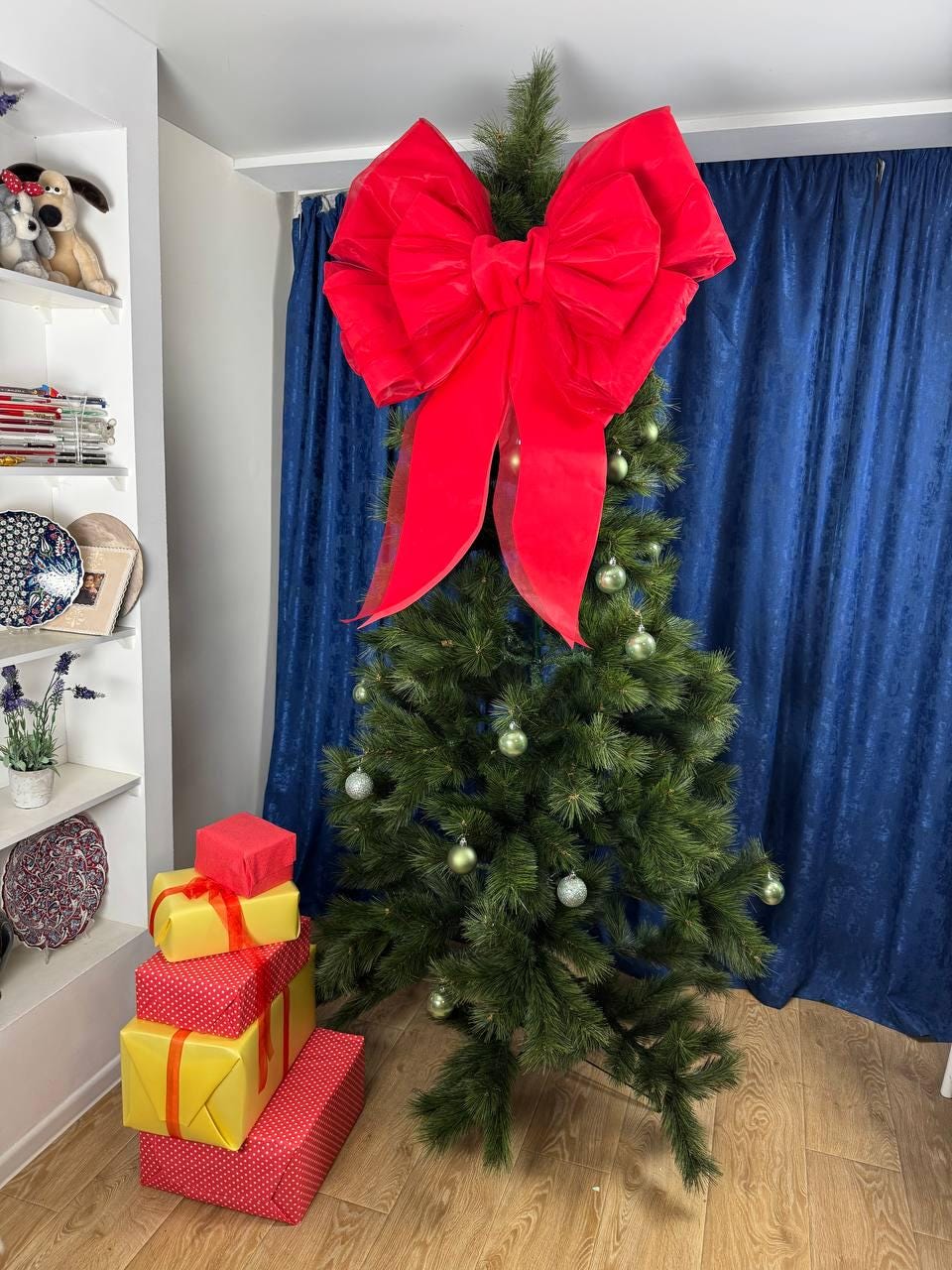 May include: A decorated Christmas tree with a large red bow on top. The tree is adorned with green ornaments. A stack of wrapped gifts in red and yellow with ribbons sits at the base of the tree. The background features a blue curtain.