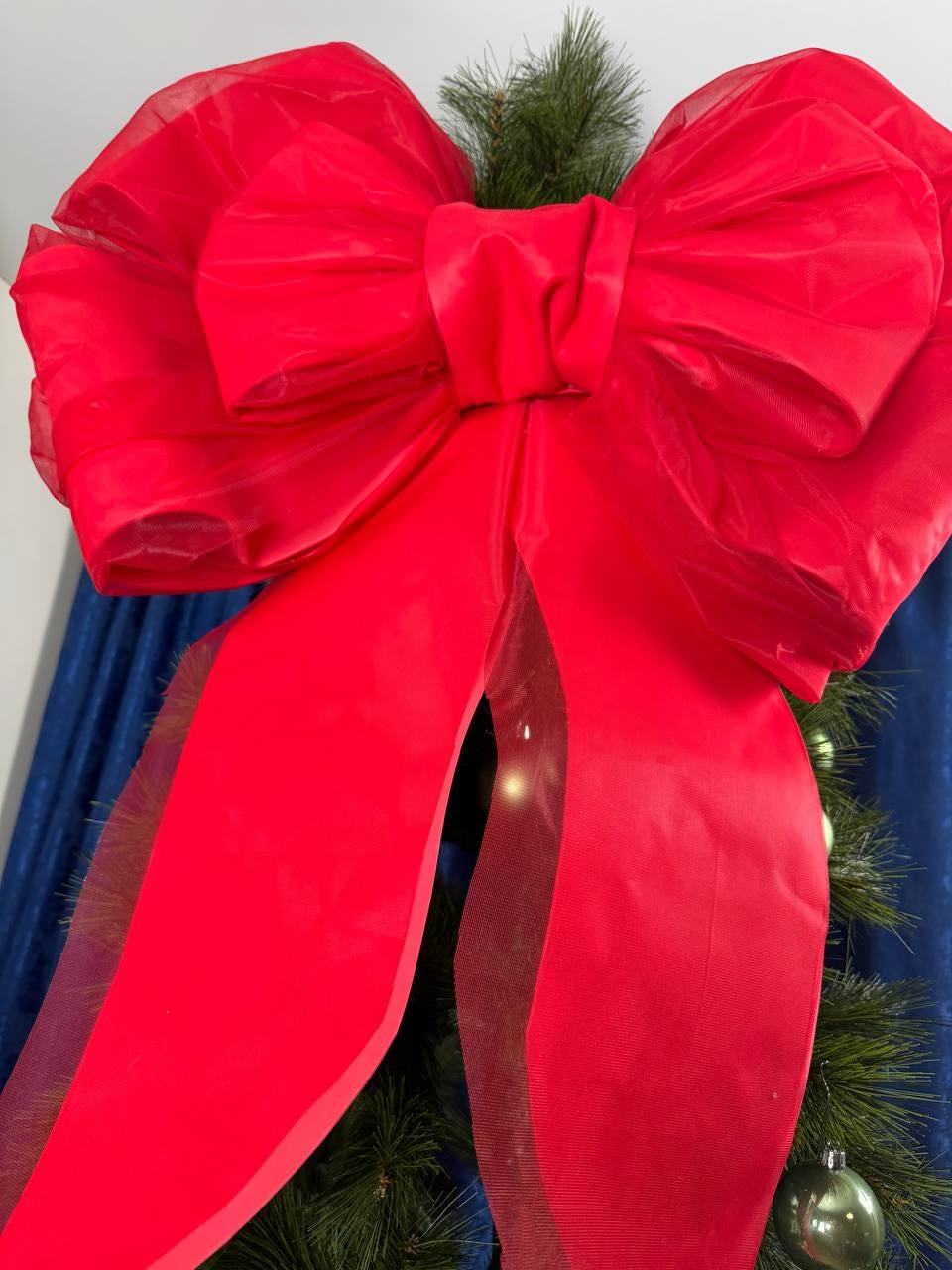 May include: A large, vibrant red bow made of a shiny fabric, possibly satin or organza, is the focal point. The bow is attached to a green Christmas tree, with a few green ornaments visible. The bow's long tails cascade downwards.