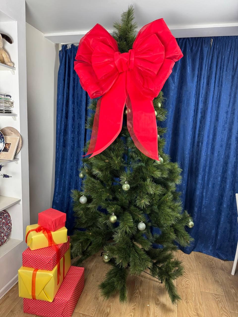 May include: A decorated Christmas tree with a large red bow at the top. The tree is adorned with silver ornaments and stands next to wrapped gifts in red and yellow. The gifts are tied with ribbons.