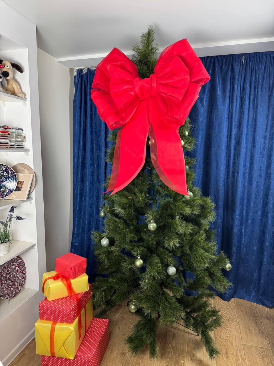 May include: A decorated Christmas tree with a large red bow at the top. The tree is adorned with silver ornaments. Several wrapped gifts in red and yellow are stacked at the base of the tree. The background includes blue curtains and a white shelf.