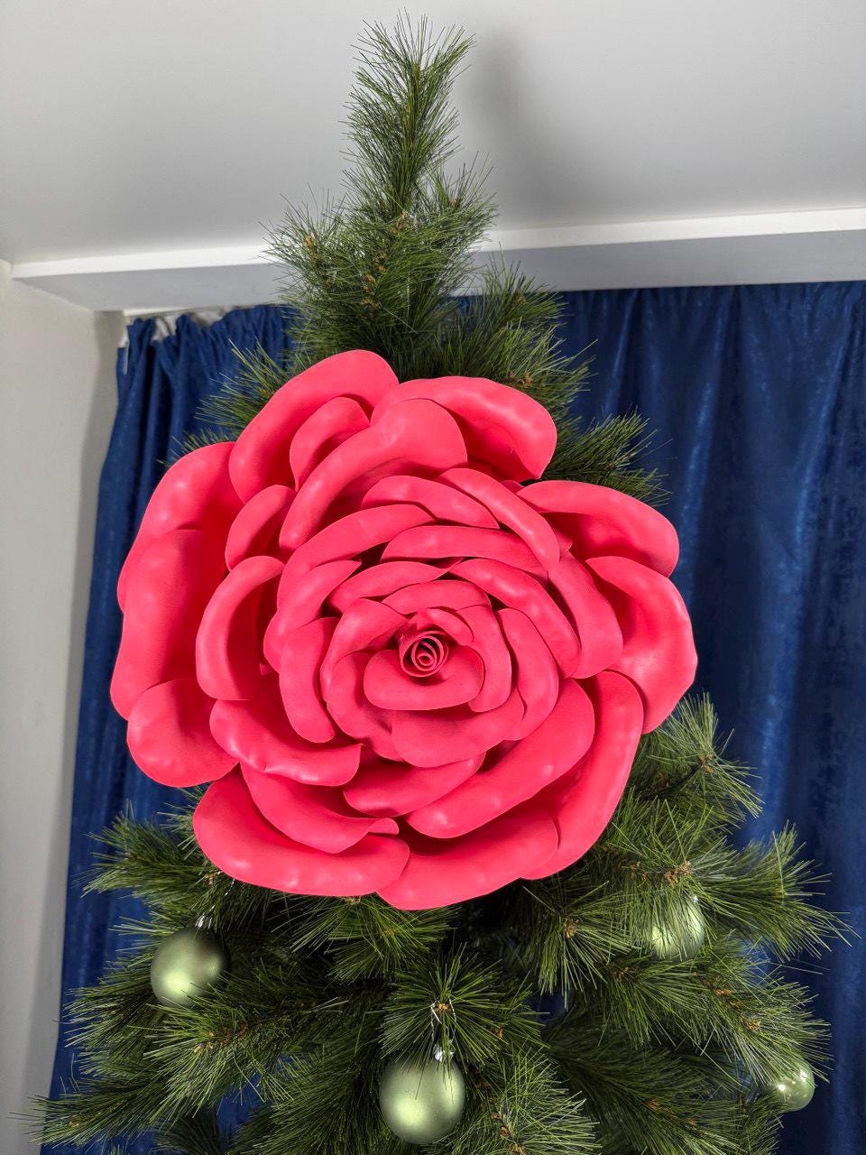May include: A large, vibrant pink rose decoration adorns a Christmas tree. The artificial rose is the focal point, surrounded by green pine needles and accented with small, round, green ornaments. The background features a blue curtain.