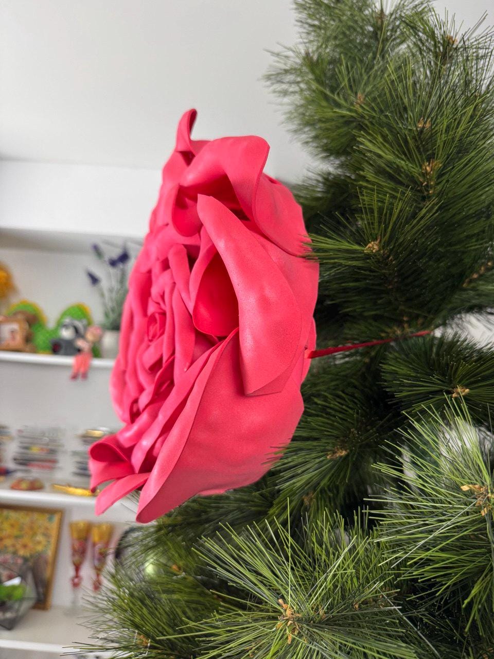 May include: A large, vibrant pink rose decoration. The artificial flower is attached to a green pine branch, creating a striking contrast. The rose's petals are detailed and textured, suggesting a handmade craft item.
