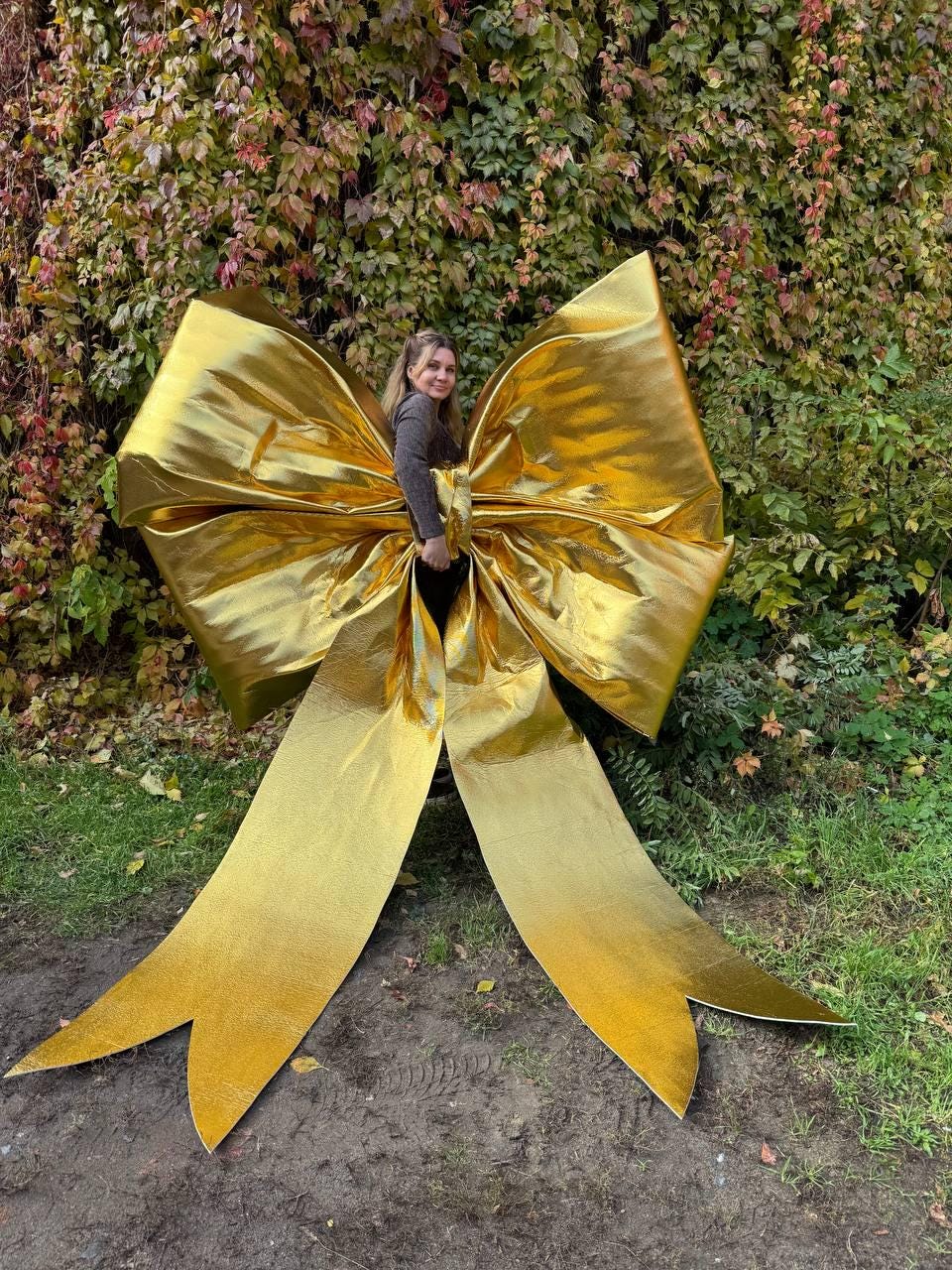 May include: A large, golden bow made of shiny material. The bow is positioned outdoors against a backdrop of green and red foliage. The bow's size suggests it could be used for a large gift or decoration.