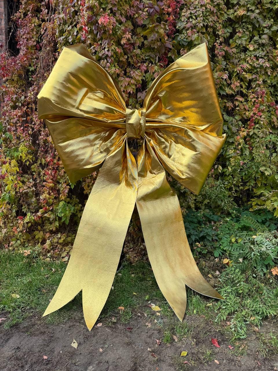 May include: A large, golden bow with long, flowing ribbons. The bow is made of a shiny, metallic material and is displayed outdoors against a backdrop of green and red foliage. The bow's ribbons have a distinctive, pointed cut at the ends.